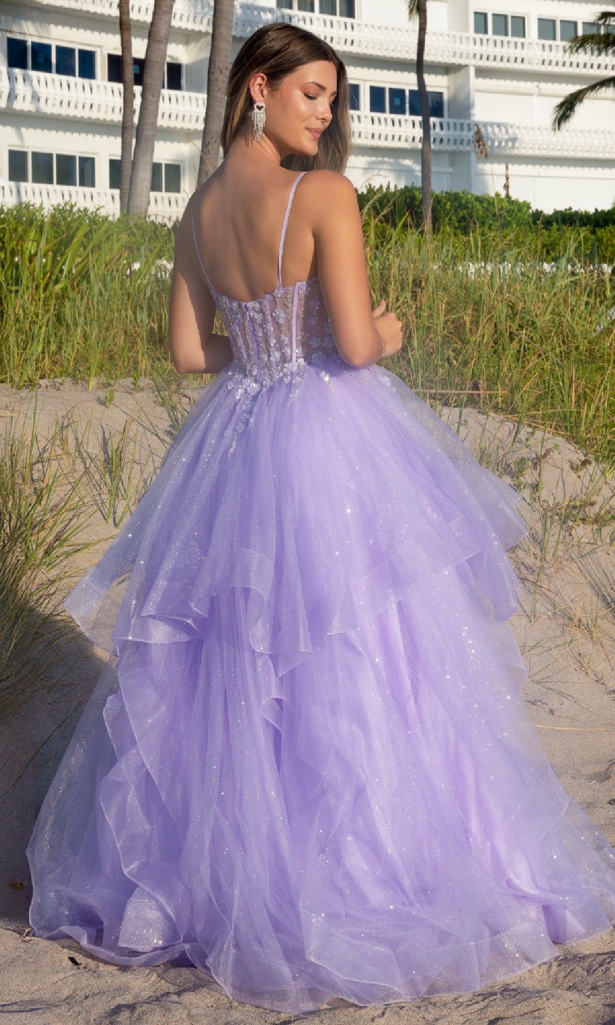 A woman stands on a sandy beach, facing away from the camera, wearing the Amelia Couture 5068 Long Glitter Prom Ball Gown—a lavender tiered tulle dress with spaghetti straps and a corset-style bodice.